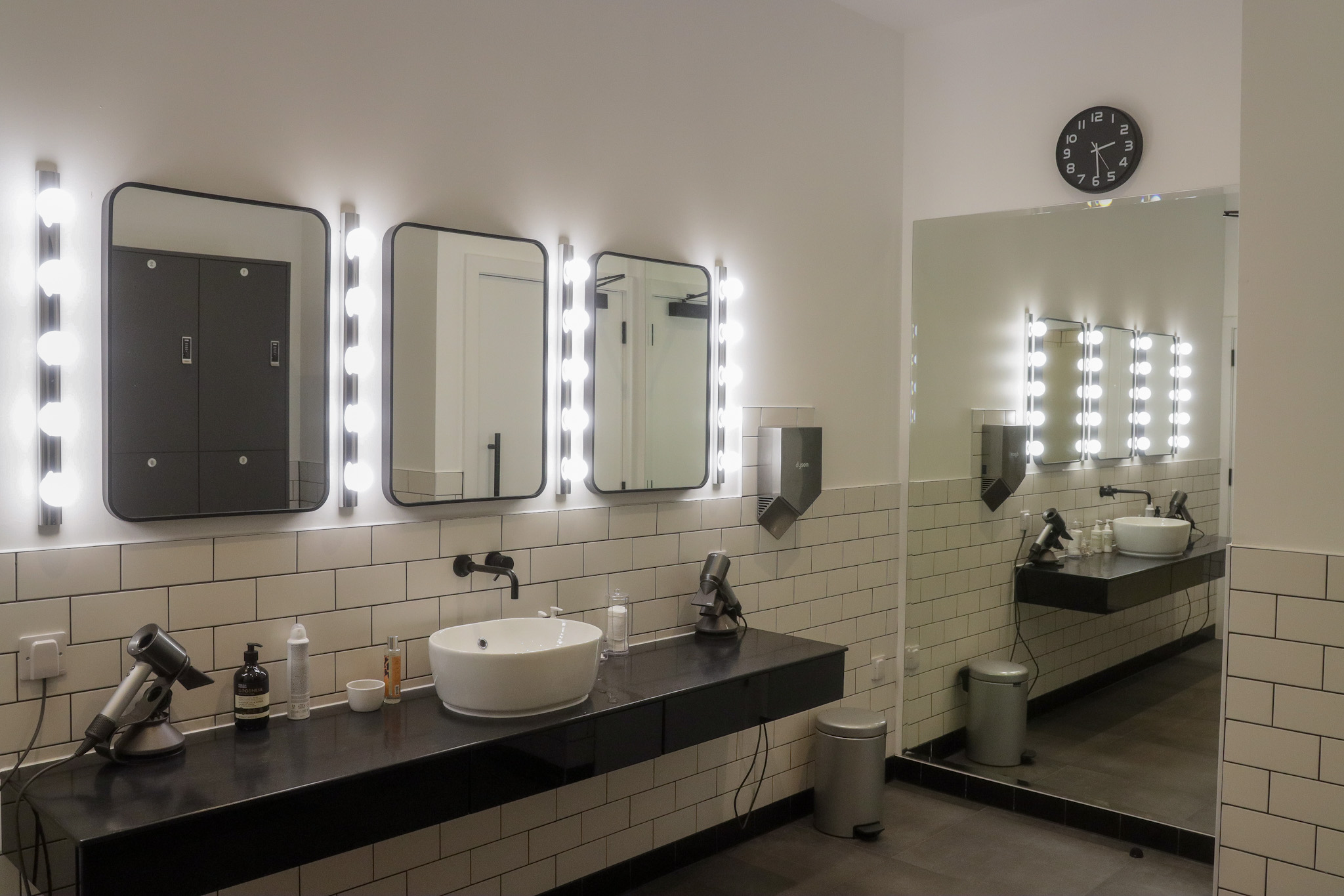 A line of mirrors with lights around them in a changing room with a large mirror on the wall to the right.