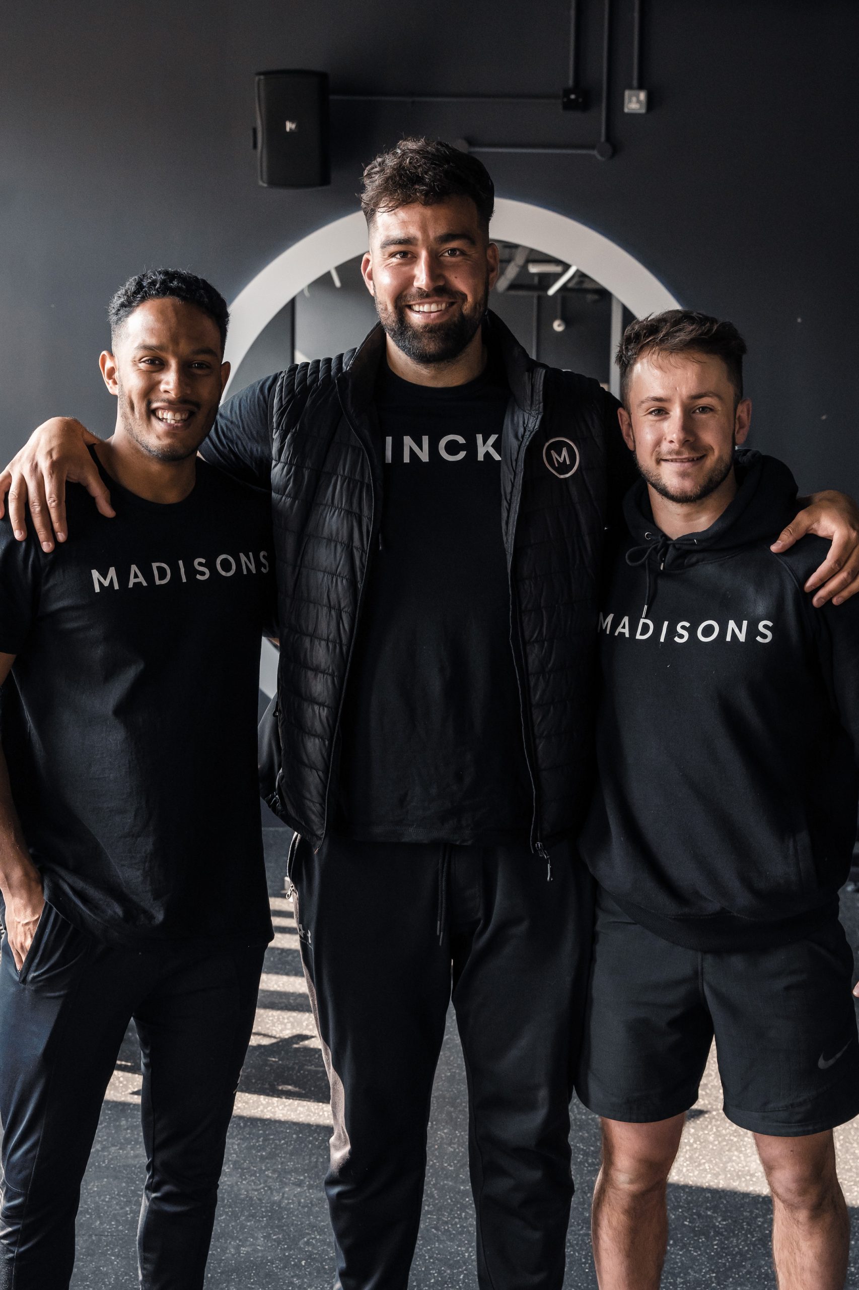Three people wearing black Madisons uniforms smiling for the camera. The person in the middle has his arms aorund the other two.