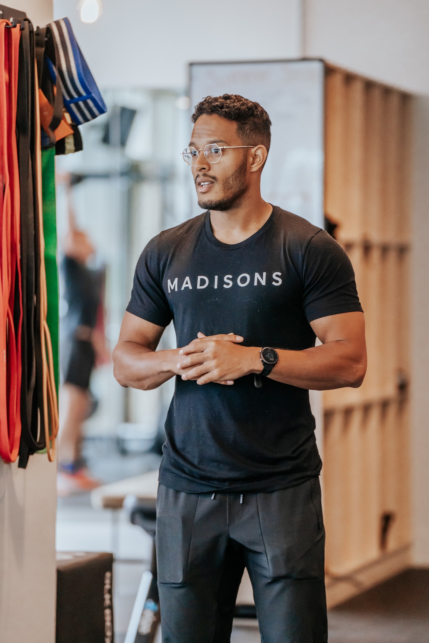 A person wearing a black t-shirt with a Madisons logo on it standing with resistance bands hanging on the wall to his left.