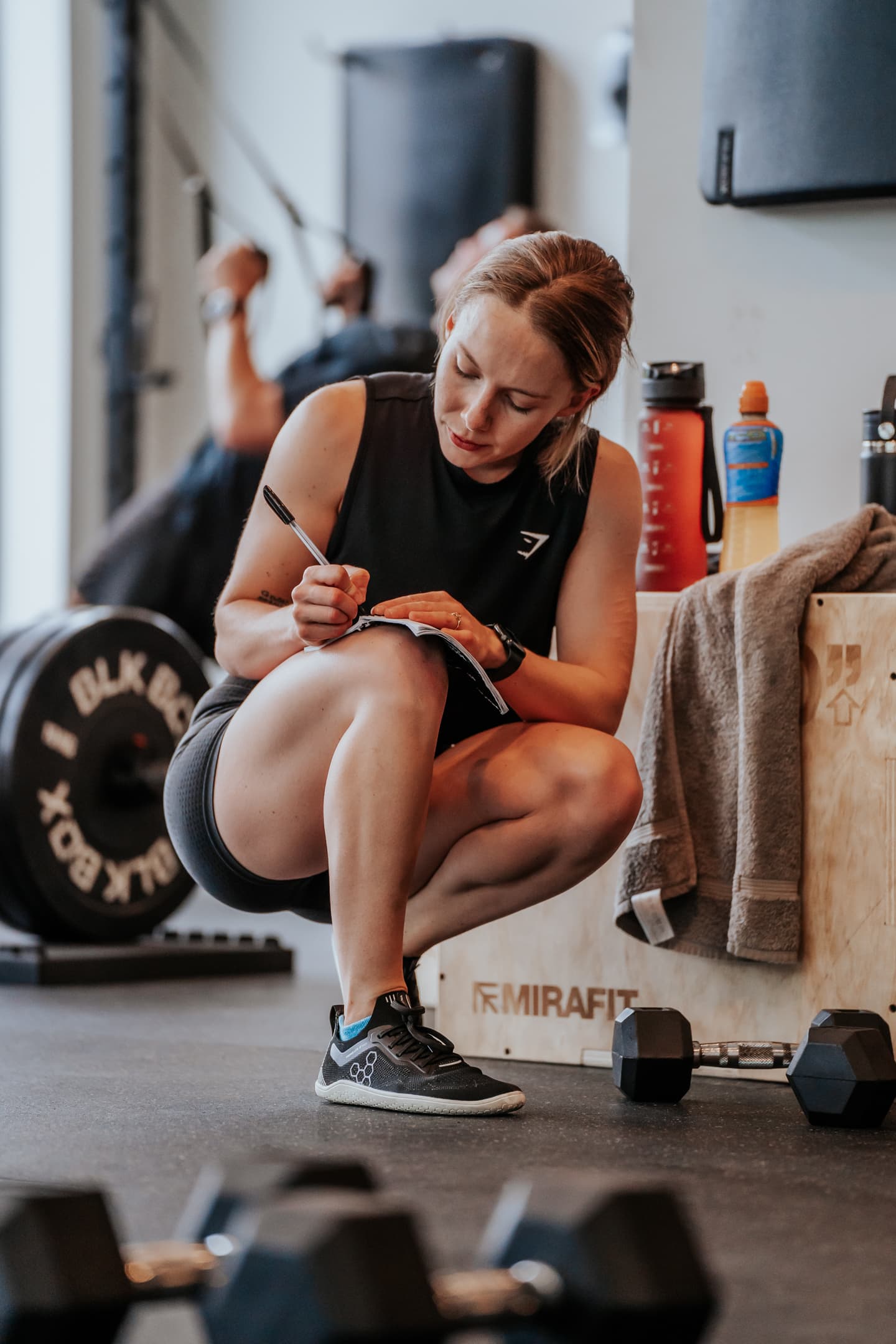 A person kneeling down writing in a notebook resting on her leg, with dumbbells in the foreground.